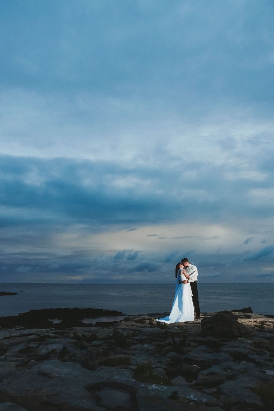 Wedding couple kissing on the rocky shore of Doolin Bay, evening clouds in the background and dramatic flash from the right lighting up the scene