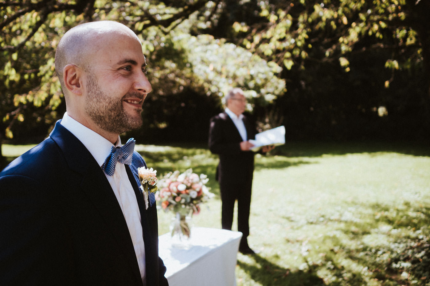 Bräutigam schaut erwartungsvoll auf die einlaufende Braut - im Hintergrund steht der Trauredner - die freie Trauung im Weingut Weidenmühle kann losgehen