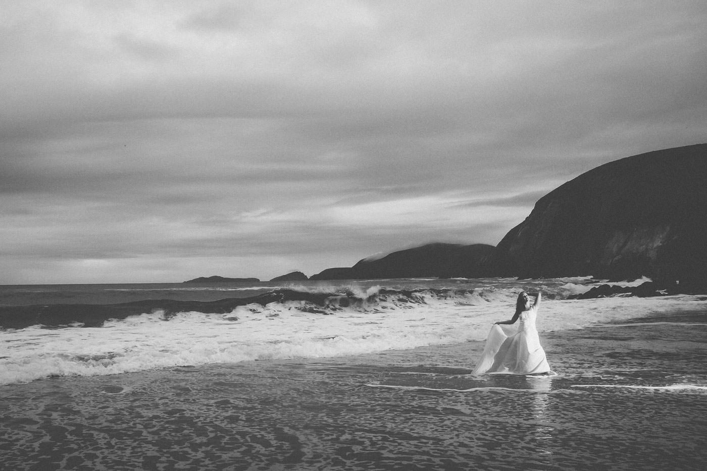Braut steht weit entfernt mit ihrem Brautkleid Wasser am Atlantik, Schwarzweissfoto, im Hintergrund Brandung und schroffe Felsen über die sich Wolken schieben, dramatische Stimmung, After-Wedding-Session am Slea Head, Dingle, Co. Kerry, Irland