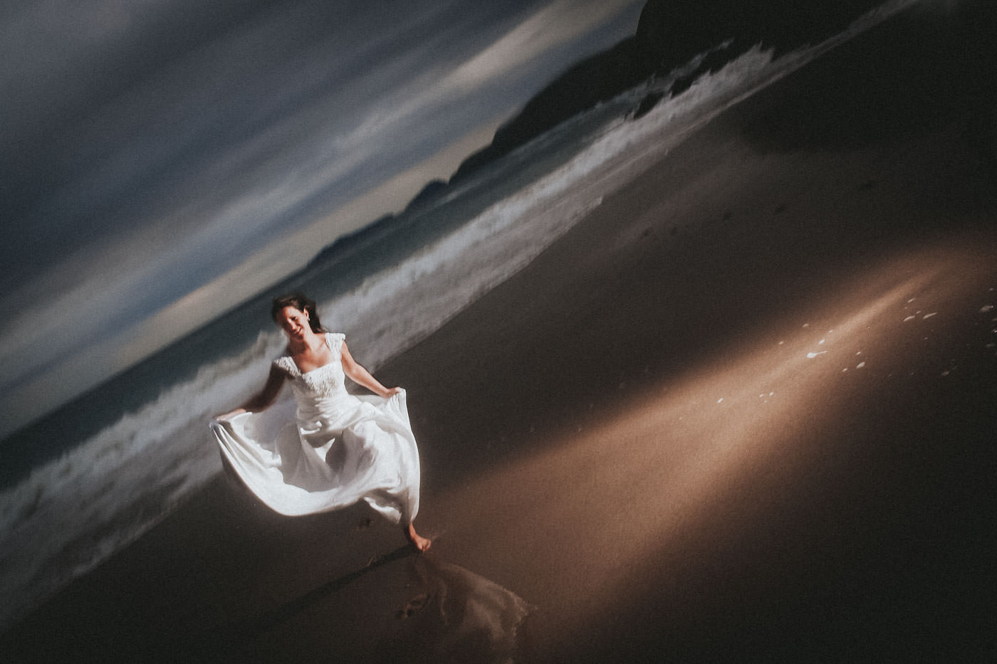 Bride with wedding dress at an Irish beach, running towards the photographer, evening light and dramatic flash, at Slea Head, Dingle, Co. Kerry, Ireland