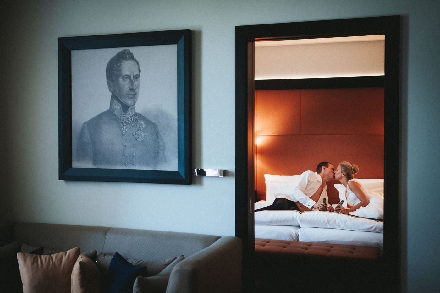 Bride and groom lying on bed in hotel room kissing, his shirt open, both with beer bottle in hand, Steigenberger Hotel Bad Homburg