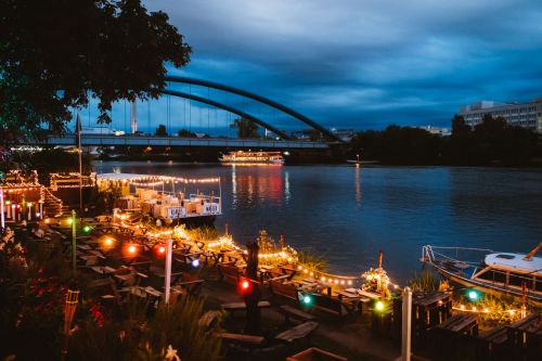 Abendliche Szene im Blaues Wasser Frankfurt, mit Blick über den erleuchteten Beach-Bereich mit Main und Schiffen im Hintergrund