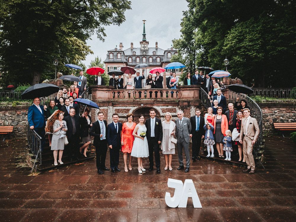 Gruppenfoto im Regen auf einer Hochzeit in Frankfurt-Höchst - Gäste mit Regenschirmen und Bolongaropalast im Hintergrund - Hochzeitsfotograf Frankfurt Brautrausch