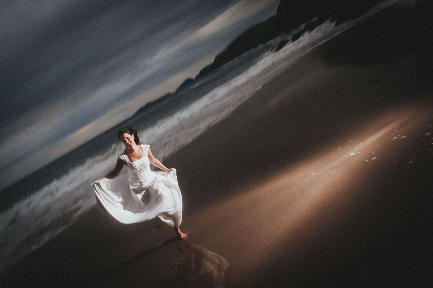 Bride with wedding dress at an Irish beach, running towards the photographer, evening light and dramatic flash, at Slea Head, Dingle, Co. Kerry, Ireland