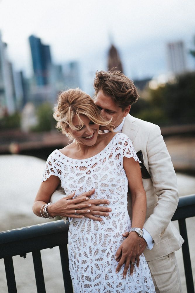 Braut im schicken Boho-Kleid im Arm des Bräutigams auf Mainbrücke vor Frankfurter Skyline - Hochzeitsfotograf Frankfurt Brautrausch