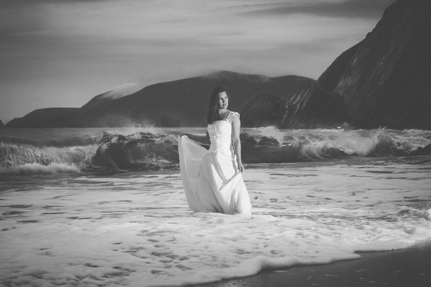 Braut mit entschlossenem Gesichtsausdruck steht mit ihrem Brautkleid Wasser am Atlantik, Schwarzweissfoto, im Hintergrund Brandung und schroffe Felsen über die sich Wolken schieben, dramatische Stimmung, After-Wedding-Session am Slea Head, Dingle, Co. Kerry, Irland