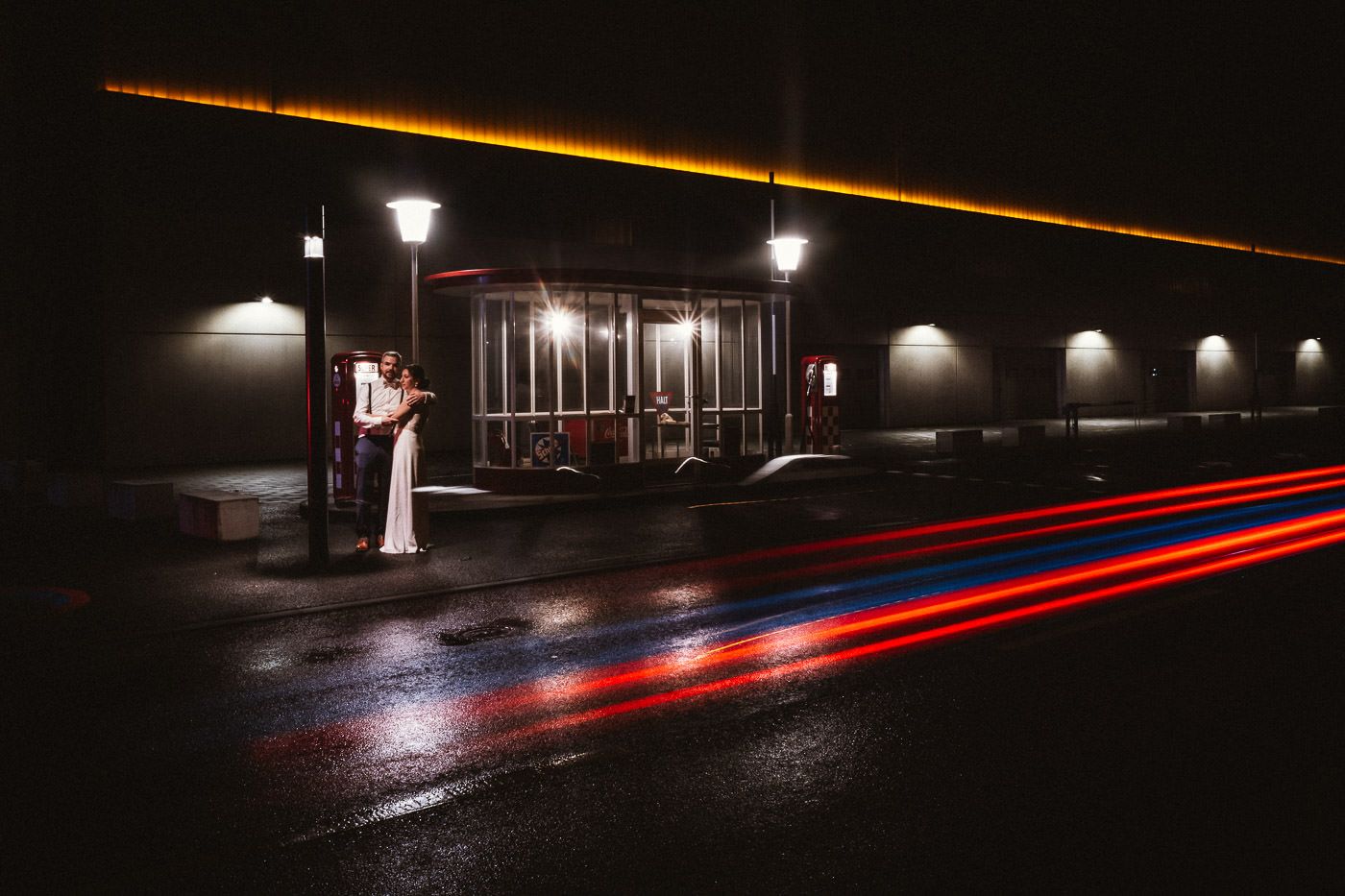 Brautpaar kuschelt bei Nacht vor historischer Tankstelle. Auf der Straße spiegeln sich die Lichter eines vorbeifahrenden Autos. Hochzeitsfotos in Frankfurt am Main.